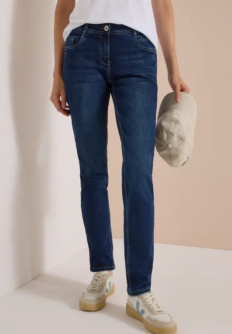 Mid Waist Slim Leg Jeans w stylu Casual Fit Dark blue wash Osoba ma na sobie ciemnoniebieskie dżinsy, białe trampki i trzyma jasną czapkę w ręku.