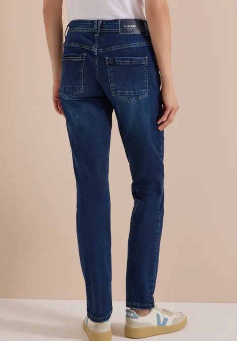 Mid Waist Slim Leg Jeans w stylu Casual Fit Dark blue wash Kobieta od tyłu w ciemnoniebieskich prostych dżinsach i jasnych trampkach na beżowym tle.