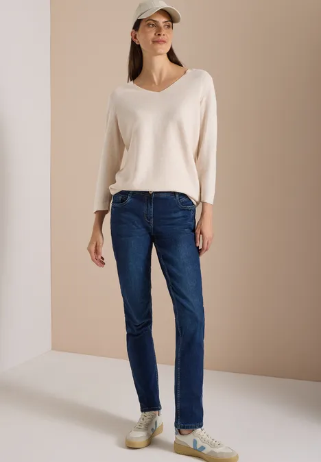 Mid Waist Slim Leg Jeans w stylu Casual Fit Dark blue wash Kobieta w ciemnoniebieskich jeansach, beżowym swetrze z dekoltem w serek i czapce na neutralnym tle.