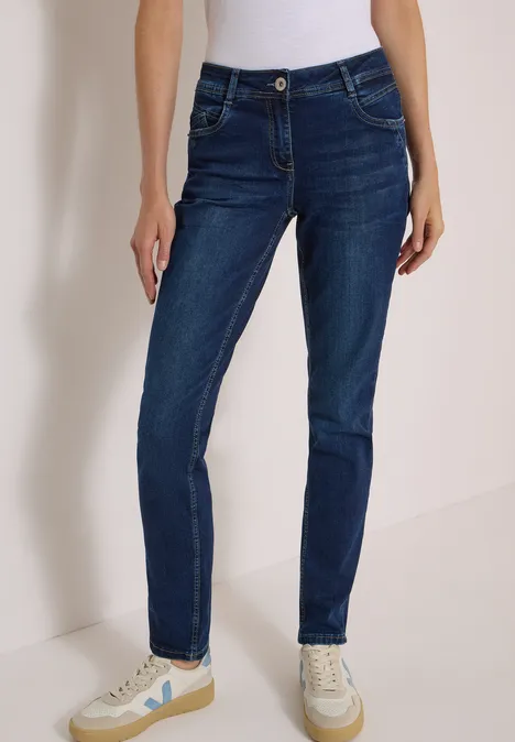 Mid Waist Slim Leg Jeans w stylu Casual Fit Dark blue wash Osoba w ciemnoniebieskich dopasowanych jeansach i jasnych trampkach na neutralnym tle.