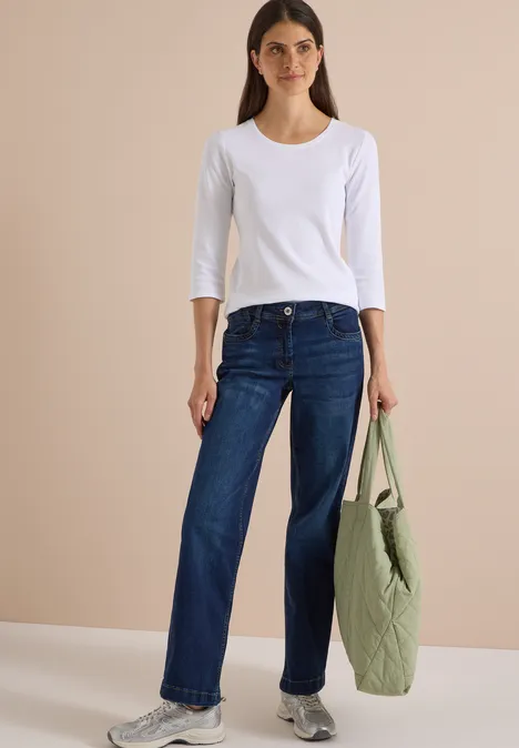 High Waist Wide Leg Jeans w kroju Loose Fit Dark blue wash Kobieta w białej bluzce z rękawami 3/4, w szerokich dżinsach trzyma zieloną torbę.