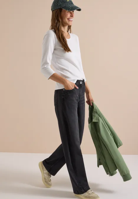 Frau in schwarzer Jeans, weißem Langarmshirt und Cap hält grüne Jacke lässig über den Arm.
