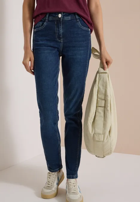 Frau trägt dunkelblaue Jeans, weiße Sneakers und hält eine beige Tasche in der Hand.