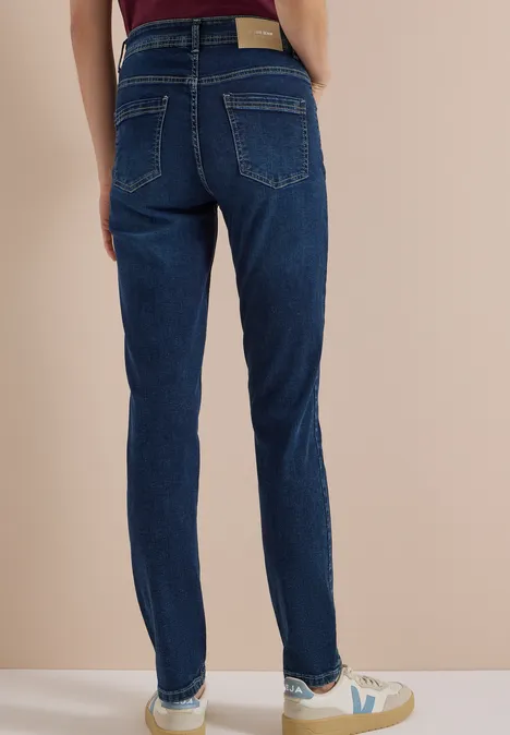 Person von hinten in dunkelblauen Slim-Fit-Jeans und weißen Sneakern vor beigem Hintergrund.