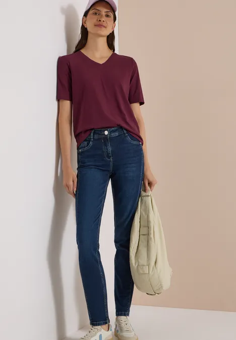 Junge Frau in lila V-Shirt, Jeans und Cap lehnt an Wand und hält eine helle Stofftasche.