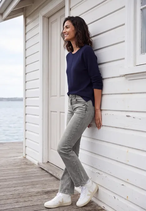 High Waist Bootcut Jeans im Slim Fit light grey wash Frau in Jeans und Pullover lehnt lächelnd an weißer Holzfassade am Wasser.
