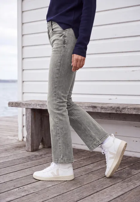 High Waist Bootcut Jeans im Slim Fit light grey wash Frau trägt hellgraue Jeans und weiße Sneaker auf einer Holzterrasse vor weißer Fassade am Wasser.