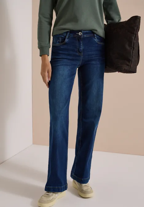 Persoon draagt donkerblauwe uitlopende jeans, groen shirt met lange mouwen en houdt een donker kussen vast.