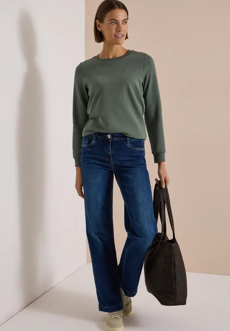 Vrouw in groene trui, blauwe jeans en lichte schoenen met zwarte tas voor een beige muur.