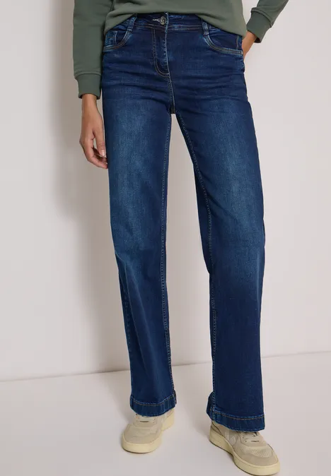 Persoon draagt donkerblauwe, rechte jeans en lichte sneakers voor een neutrale achtergrond.