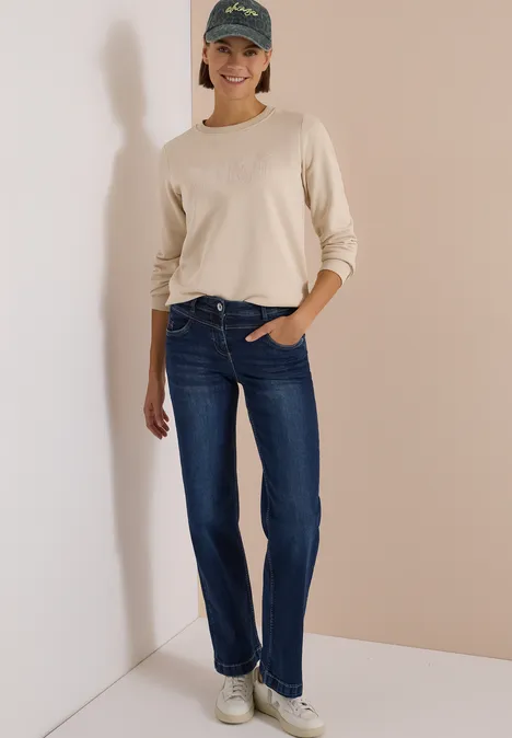 Een lachende vrouw in jeans, beige shirt, witte sneakers en pet staat tegen een muur.