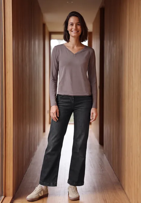 High Waist Wide Leg Jeans im Slim Fit black washed Eine Frau steht lächelnd in einem holzvertäfelten Flur, trägt dunkle Jeans und langärmeliges Oberteil.