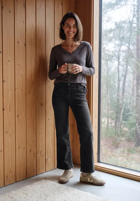 High Waist Wide Leg Jeans im Slim Fit black washed Eine lächelnde Frau steht in einem Holzhaus neben einem Fenster und hält eine Tasse.