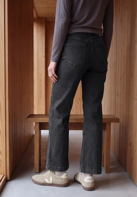 High Waist Wide Leg Jeans im Slim Fit black washed Eine Person von hinten trägt schwarze Jeans und beige Sneaker, steht in einem hellen Holzraum.