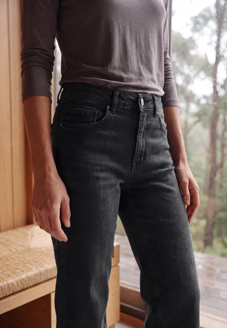 High Waist Wide Leg Jeans im Slim Fit black washed Frau in grauem Langarmshirt und schwarzen Highwaist-Jeans, stehend vor Fenster mit Waldsicht.