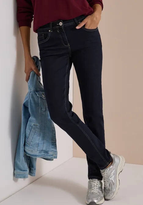 Mid Waist Slim Leg Jeans im Casual Fit rinsed wash Model lehnt an Wand, trägt dunkelblaue Jeans, silberne Sneakers und hält eine hellblaue Jeansjacke.