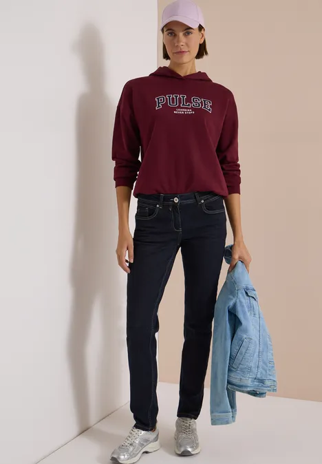 Mid Waist Slim Leg Jeans im Casual Fit rinsed wash Frau in burgunderrotem Pullover, Jeans und Cap, hält eine Jeansjacke, trägt silberne Sneaker im Minimal-Setting.