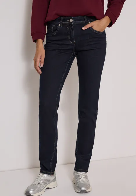 Mid Waist Slim Leg Jeans im Casual Fit rinsed wash Frau trägt dunkelblaue Jeans und helle Sneaker, posiert vor weißem Hintergrund.