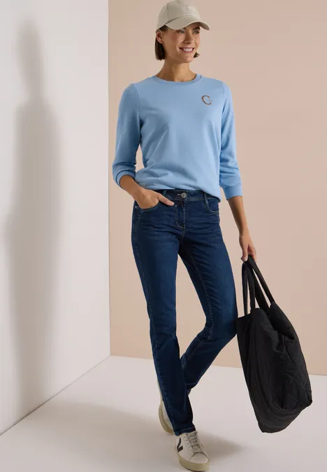 Vrouw in lichtblauw shirt, donkere jeans, witte pet en grote zwarte tas lacht ontspannen.