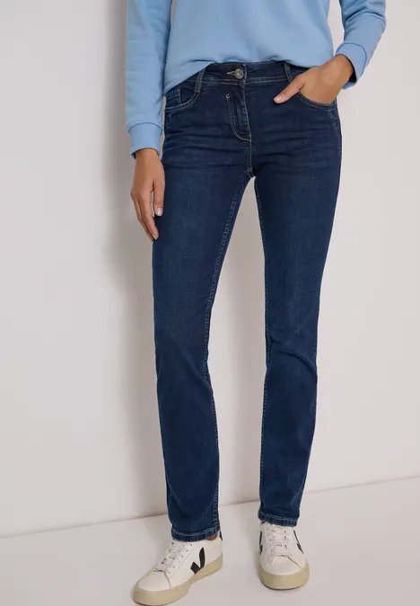 Vrouw met donkerblauwe slim-fit jeans, lichtblauw shirt en witte sneakers tegen neutrale achtergrond.