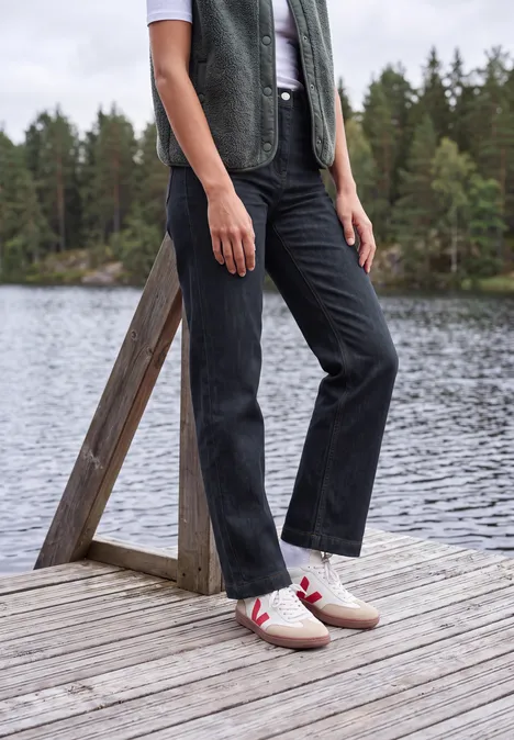 High Waist Wide Leg Jeans im Loose Fit rinsed wash Eine Person steht auf einem Holzsteg am See, trägt schwarze Hose, weiße Sneakers mit rotem Streifen und grüne Weste.
