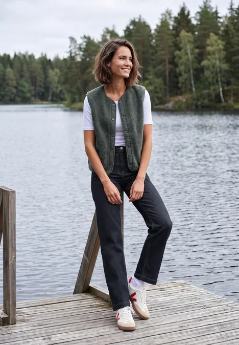 High Waist Wide Leg Jeans im Loose Fit rinsed wash Frau lehnt lächelnd an einem Steg am See, trägt Weste, T-Shirt, Jeans und Sneaker.