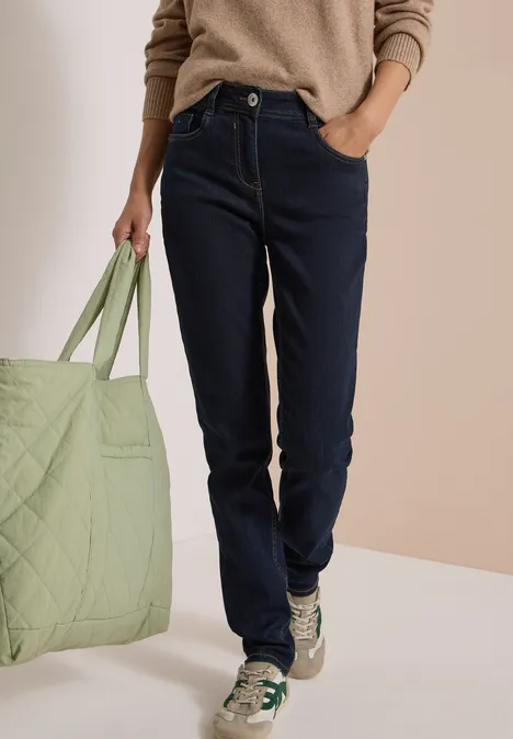 Person in dunklen Jeans, beigem Pullover und Sneakern hält eine große, hellgrüne Tasche.