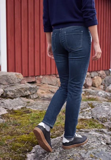 Person in Jeans und schwarzen Sneakers geht auf felsigem, moosbewachsenem Boden vor einer roten Holzwand.