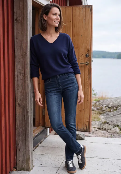 Eine Frau in Jeans und blauem Pullover lehnt lächelnd an einer Holzwand am Seeufer.