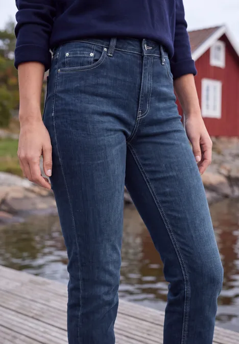 Person in dunkelblauen Jeans und blauem Pullover steht auf Steg vor rotem Haus am Wasser.