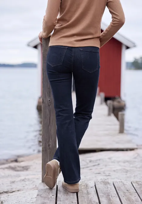 Frau in Jeans und Pullover geht über Steg zum roten Bootshaus am See.