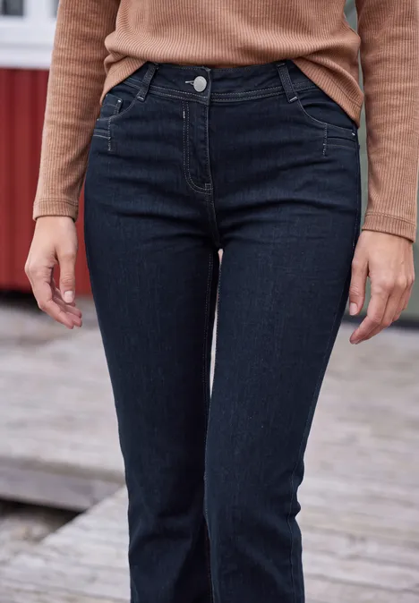 Eine Person trägt einen beigefarbenen Pullover und dunkelblaue Slim Jeans, stehend auf Holzplanken.