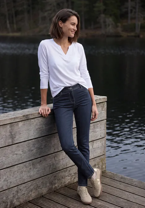 Eine Frau lehnt entspannt an einem Holzgeländer am See, trägt weiße Bluse, dunkle Hose und lächelt.