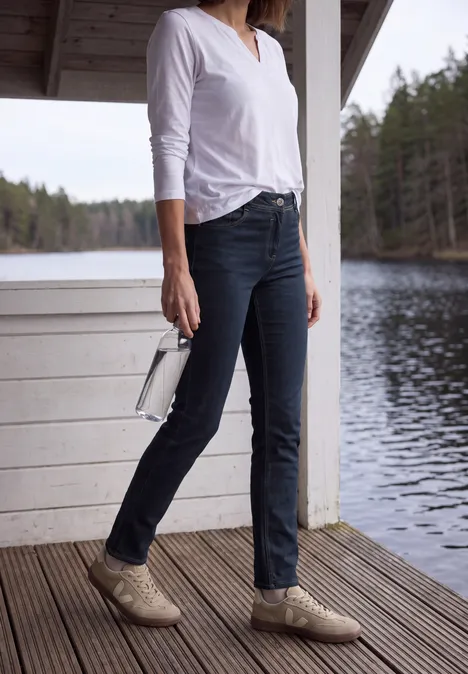 Frau in weißem Shirt und dunklen Jeans, mit beigen Schuhen und weißer Tasche, steht auf Holzdiele am See.