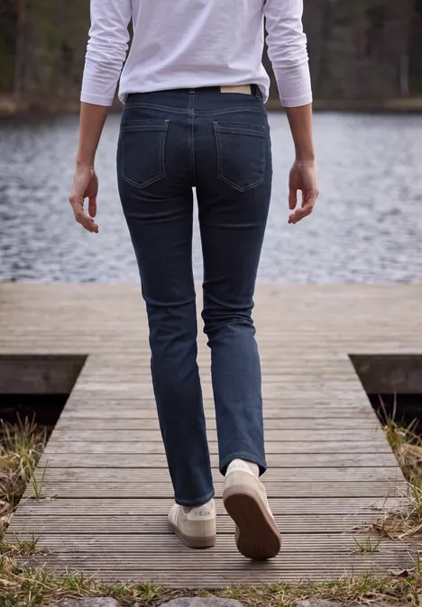 Person in engen Jeans und weißen Sneakern geht auf einem Holzsteg am See entlang.