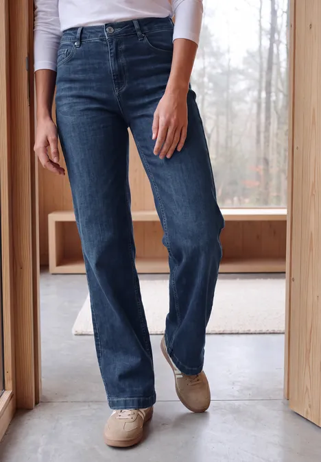 Wide Leg Jeans mit Gallonstreifen mid blue used wash Eine Person trägt dunkle, gerade geschnittene Jeans und beigefarbene Schuhe in einem hellen Raum mit Holzwänden.
