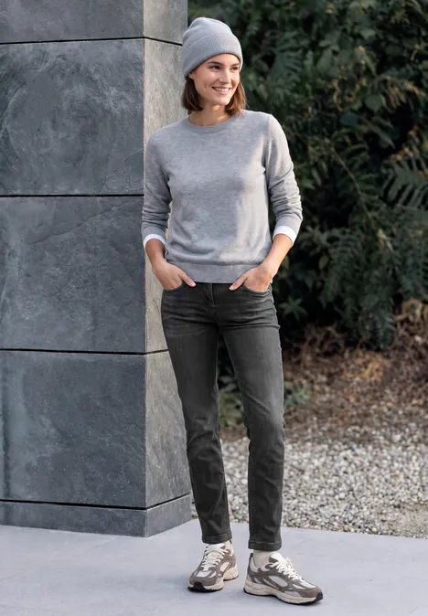 Eine Frau in grauem Pulli, Jeans und Mütze lehnt an einer Steinwand im Freien mit Pflanzen im Hintergrund.