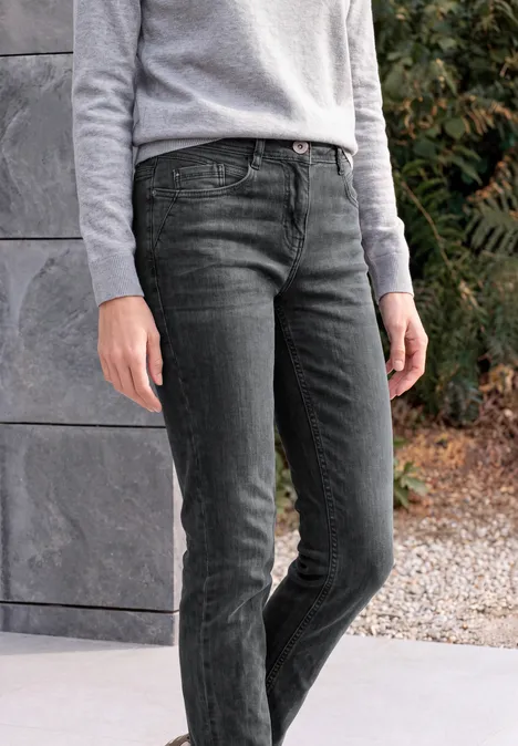 Person in grauem Oberteil und schwarzen Jeans vor einer Steinwand mit Pflanzen.