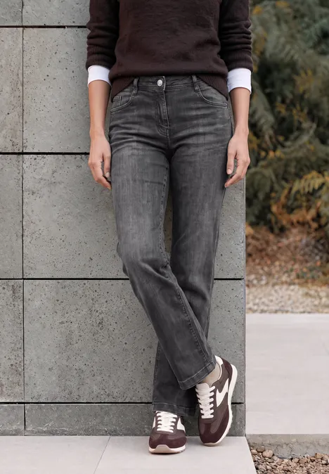 Femme adossée à un mur, jean gris foncé et baskets marron.
