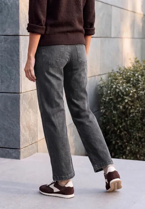 Personne de dos en pantalon gris, pull marron et baskets, devant un mur en pierre et des plantes.