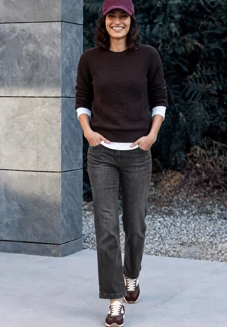 Femme décontractée avec casquette, pull marron et pantalon foncé, appuyée sur une colonne extérieure, souriante.