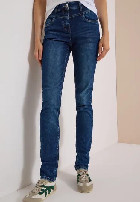 Een persoon draagt donkerblauwe skinny jeans en witte sneakers met groene details tegen een eenvoudige achtergrond.