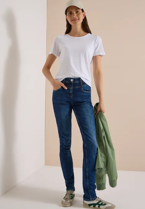 Vrouw in wit T-shirt, spijkerbroek en sneakers met pet, houdt groene jas vast voor lichte achtergrond.