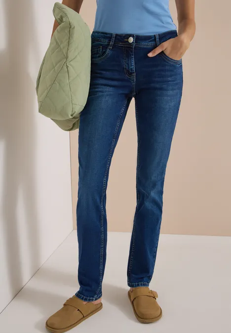 Mid Waist Straight Leg Jeans im Casual Fit mid blue used wash Person trägt blaue Jeans, hellblaues Top und braune Hausschuhe, hält eine hellgrüne Stepptasche.