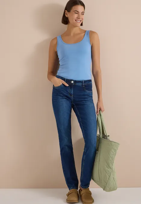 Mid Waist Straight Leg Jeans im Casual Fit mid blue used wash Frau in blauem Top und Jeans hält grünen, gesteppten Shopper vor beigem Hintergrund.