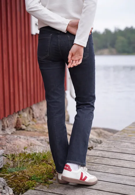 Person in dunklen Jeans und weißen Sneakern steht auf einem Steg neben einer roten Hütte am See.