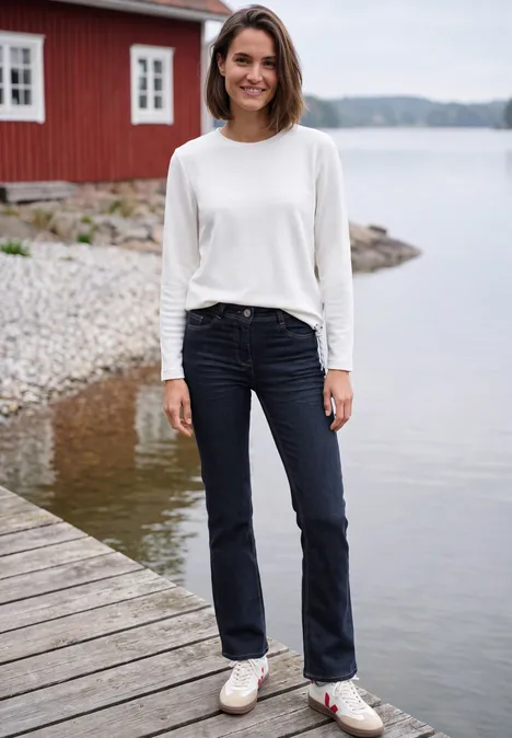 Eine lächelnde Frau in weißem Pullover und Jeans steht auf einem Holzsteg am See vor einem roten Haus.