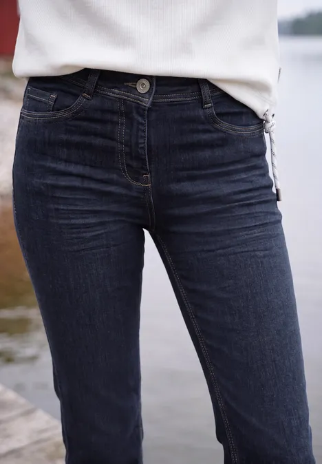 Eine Person trägt dunkelblaue Jeans und ein weißes Shirt, stehend auf einem Steg am Wasser.