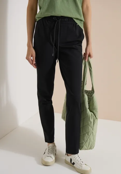 Spodnie Mid Waist z wąskimi nogawkami o Casual Fit Black Kobieta ma na sobie czarne spodnie dresowe, białe trampki, zielony top i pikowaną zieloną torbę.