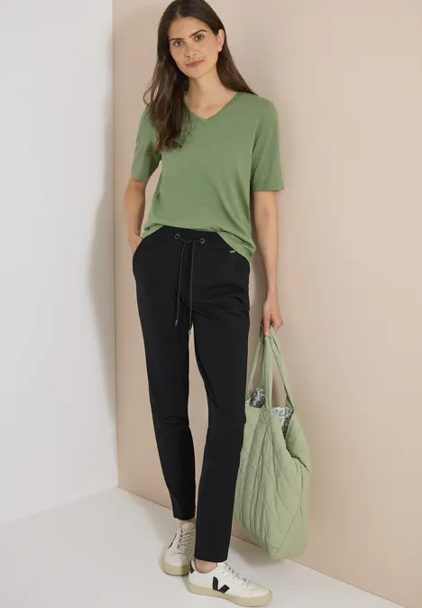 Spodnie Mid Waist z wąskimi nogawkami o Casual Fit Black Kobieta w zielonym t-shircie, czarnych spodniach dresowych i sneakersach, pozuje przed beżową ścianą z zieloną torbą.